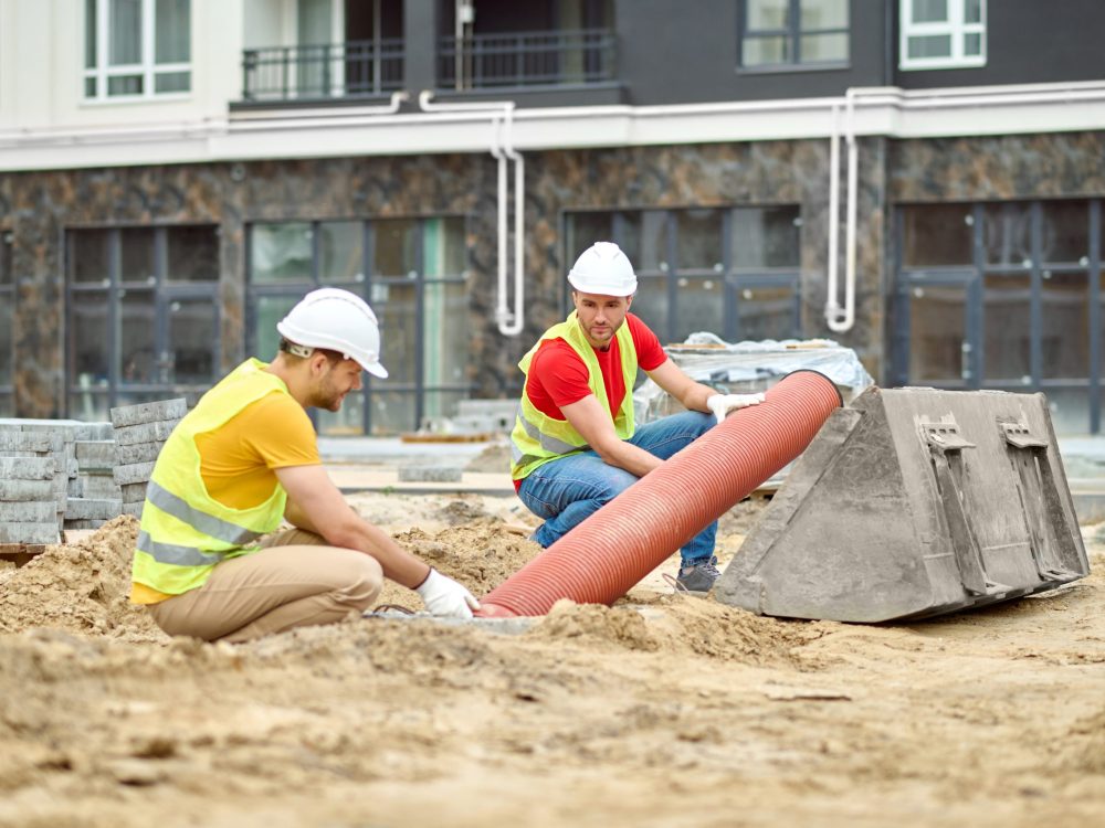 Working mood. Two men in protective helmet and bright vest crouched touching pipe on sand at construction site during day